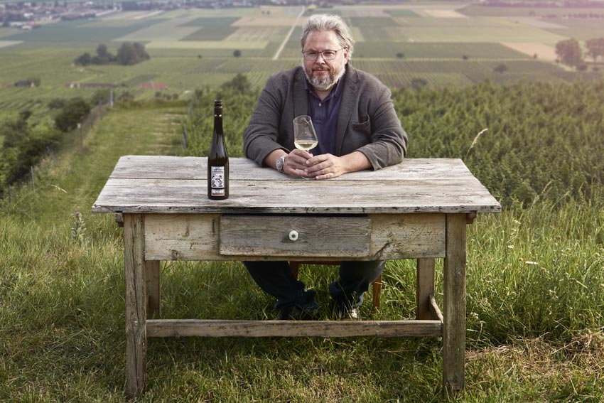 Winzer Bernhard Ott sitzt an einem Holztisch mitten im Weingarten, auf dem Tisch eine Flasche Grüner Veltliner.